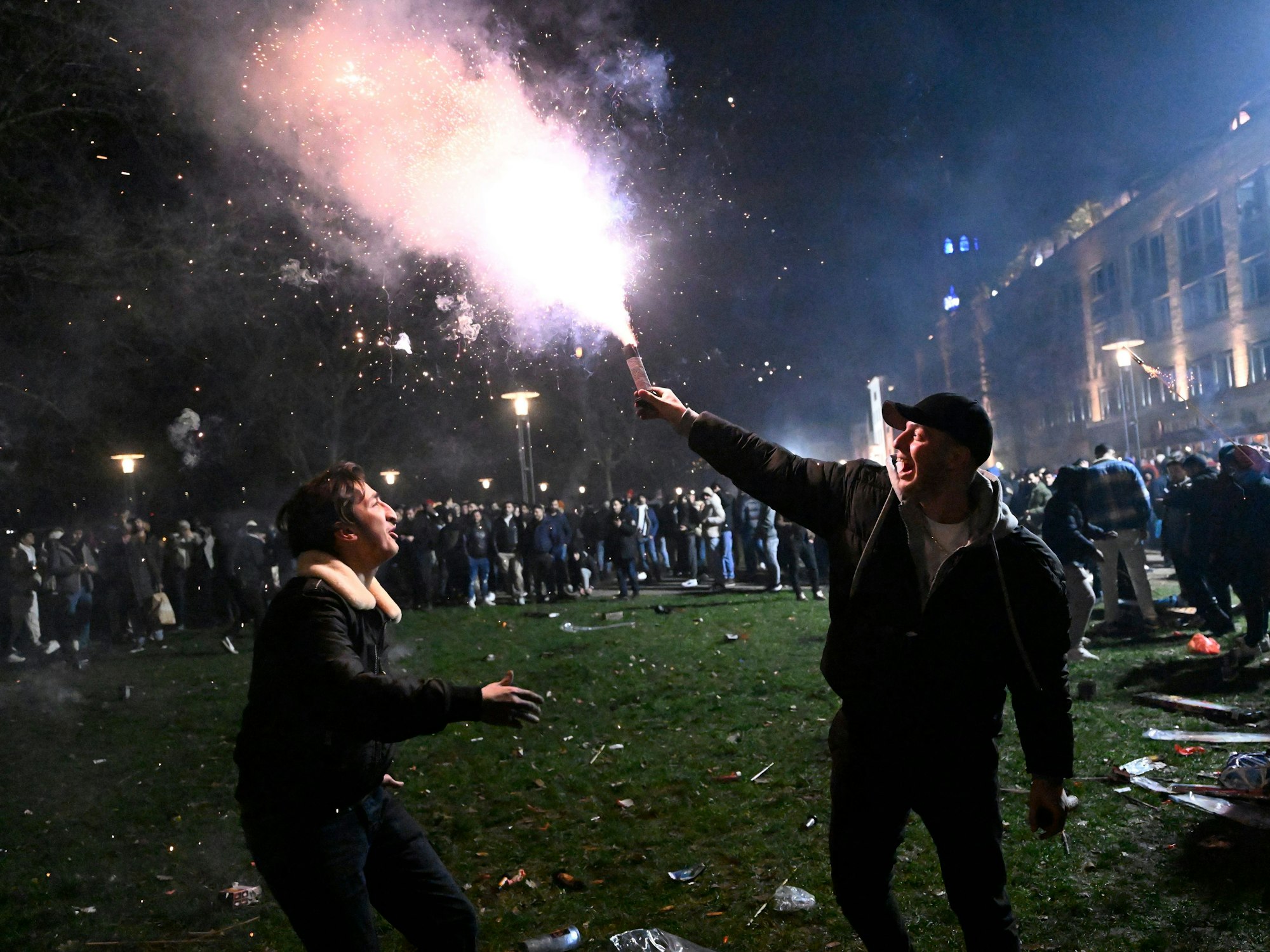 Ein junger Mann hält auf einer Wiese einen Feuerwerkskörper in der Hand und lacht einen anderen jungen Mann an.