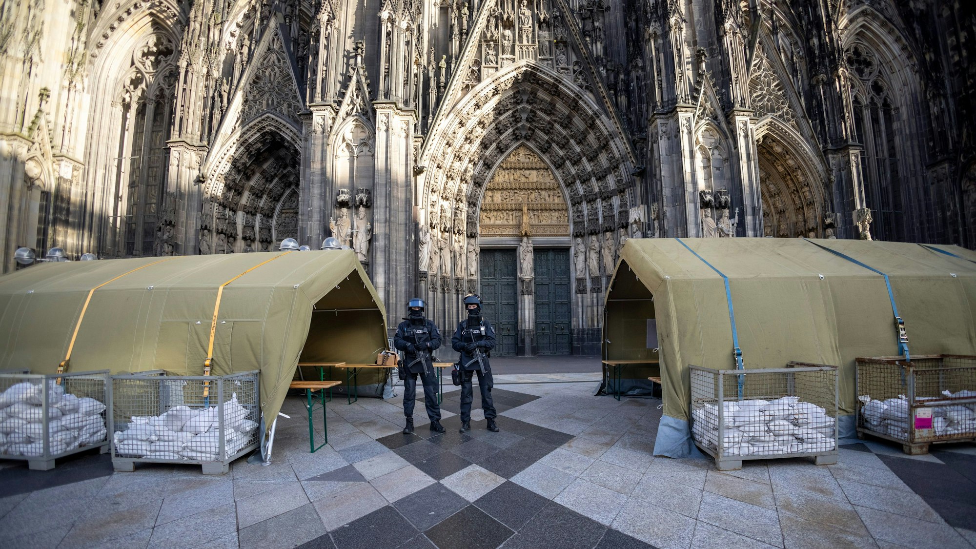 Polizisten stehen mit Maschinenpistolen vor dem Kölner Dom. Neben ihnen stehen Zelte.