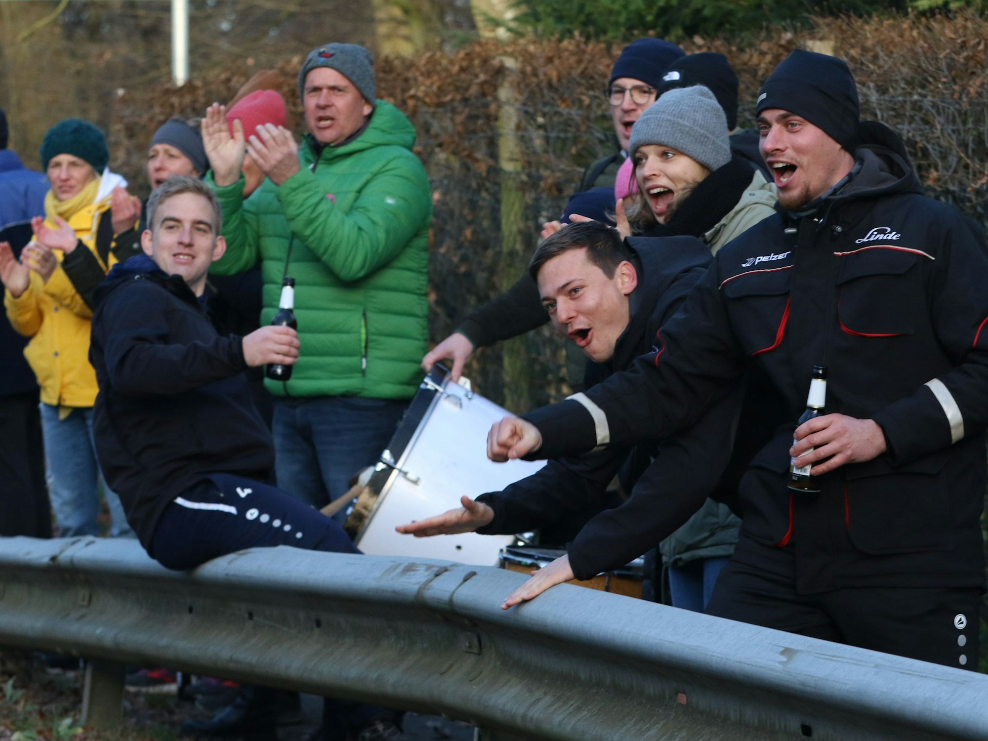 Das Bild zeigt einige Fans hinter einer Leitplanke. Sie machen Stimmung, jubeln und haben ein Bier in der Hand.