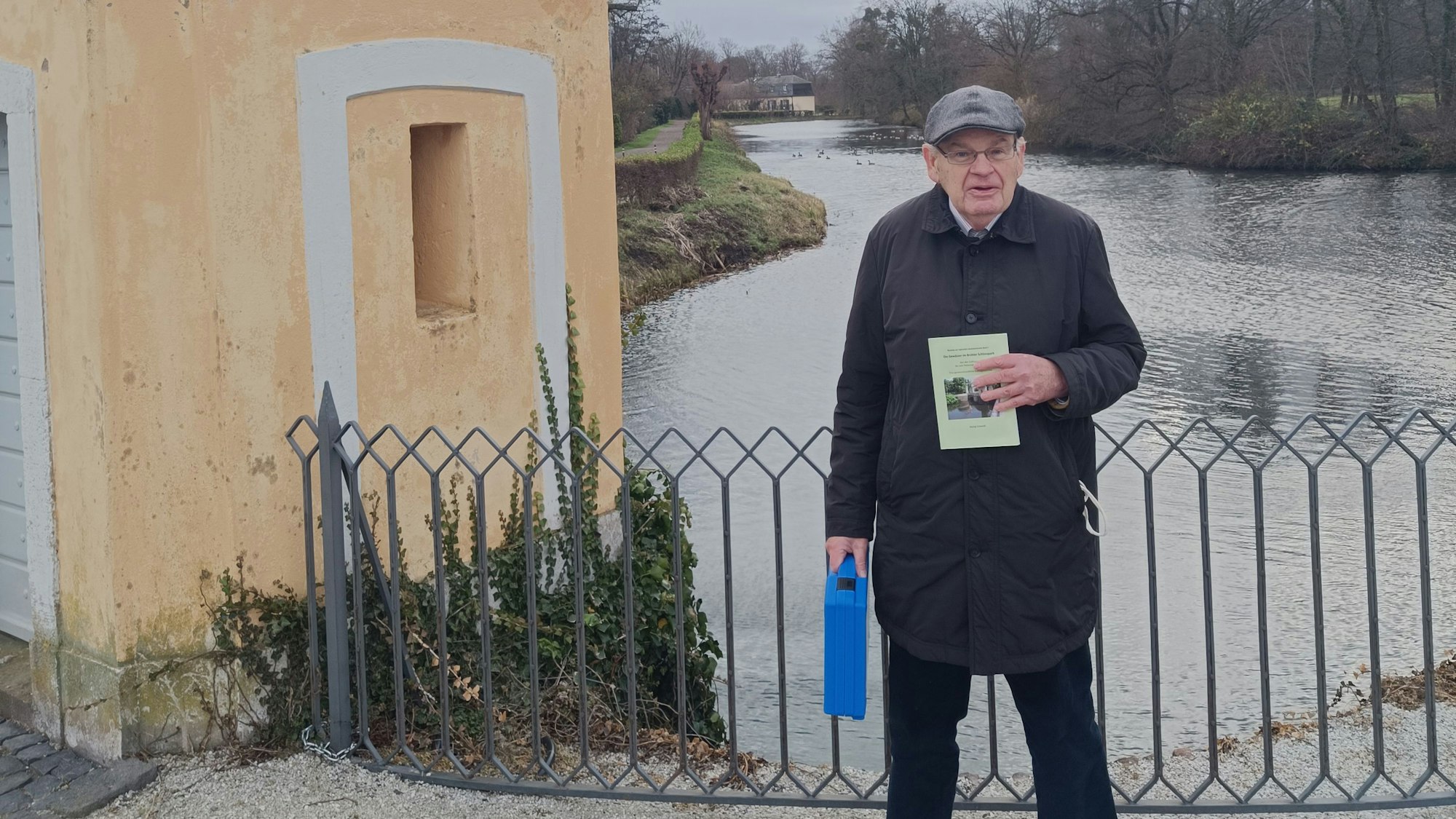Ein älterer Mann hält ein Buch in die Kamera, hinter ihm ist eines der Gewässer im Park von Schloss Augustusburg zu sehen.