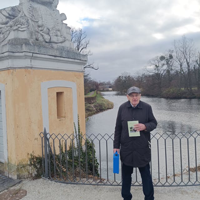 Ein älterer Mann hält ein Buch in die Kamera, hinter ihm ist eines der Gewässer im Park von Schloss Augustusburg zu sehen.&nbsp;