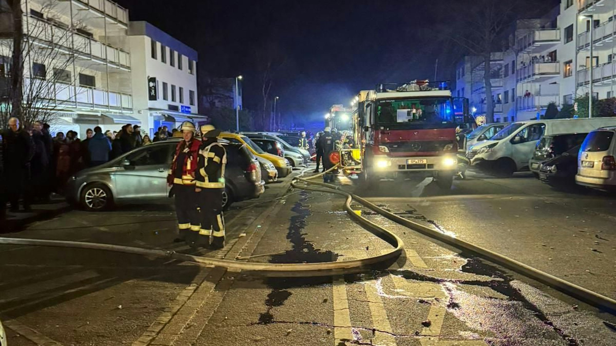 Ein Feuerwehrfahrzeug steht in der Dunkelheit auf einer Straße zwischen Mehrfamilienhäusern, Rettungskräfte und eine Menschengruppe sind zu sehen.