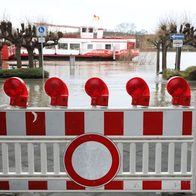 Ein „Durchfahrt verboten“-Schild und eine Absperrung stehen auf der teilweise überfluteten Rheinallee, im Hintergrund ist das Schiff „Alte Liebe“ zu erkennen.
