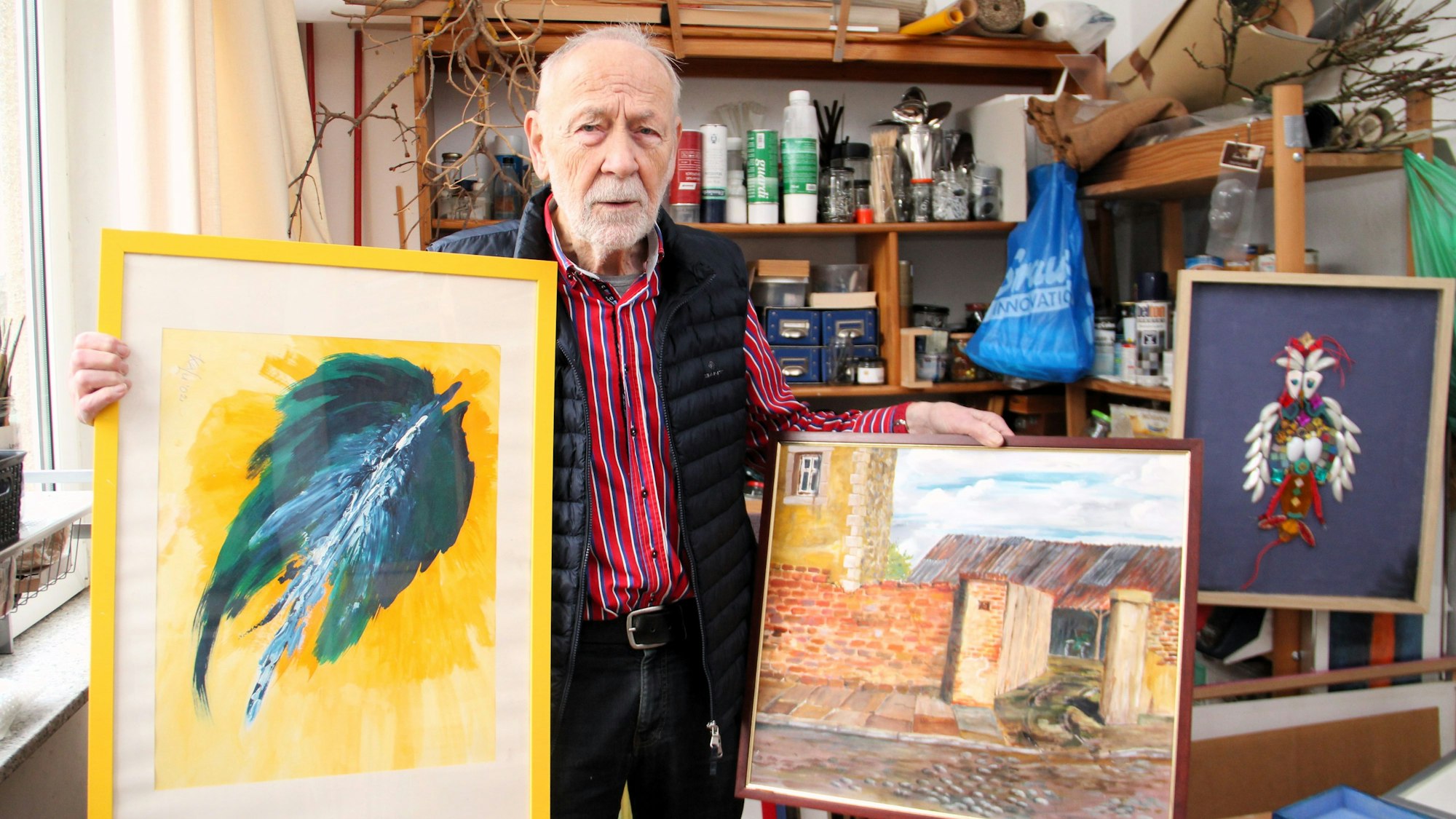 Ein älterer Mann steht in einem Atelier, er hat zwei Bilder in der Hand.