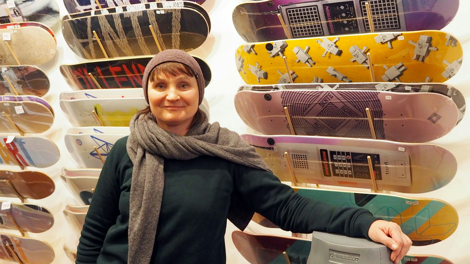 Eine Frau in Pulli, Schal und Mütze steht vor einer Wand, an der Skateboards in vielen Designs hängen.