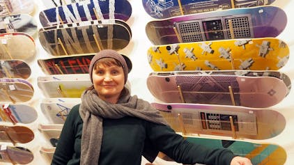 Eine Frau in Pulli, Schal und Mütze steht vor einer Wand, an der Skateboards in vielen Designs hängen.&nbsp;