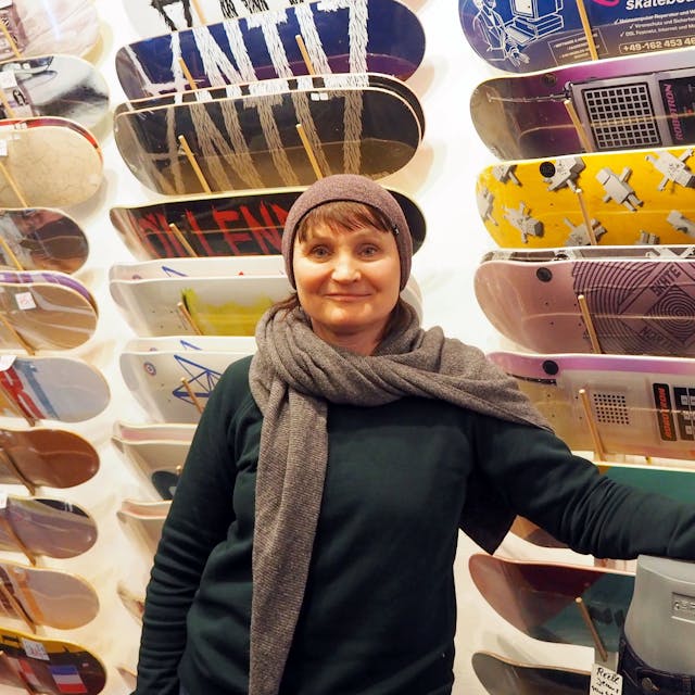 Eine Frau in Pulli, Schal und Mütze steht vor einer Wand, an der Skateboards in vielen Designs hängen.&nbsp;