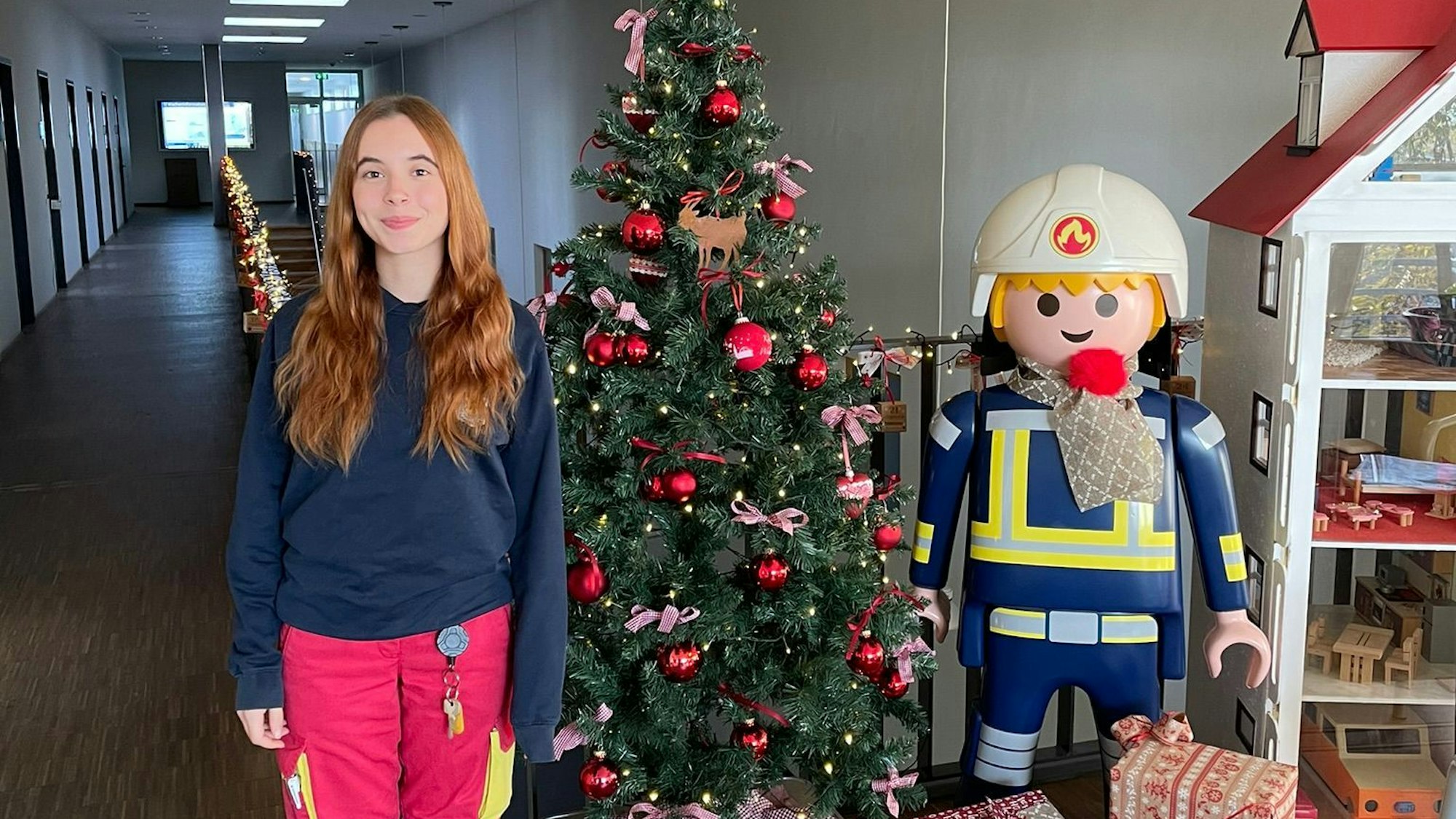 Eine junge Frau in Feuerwehruniform steht neben einem Weihnachtsbaum und einem etwa gleich großen Playmobil-Feuerwehrmann.