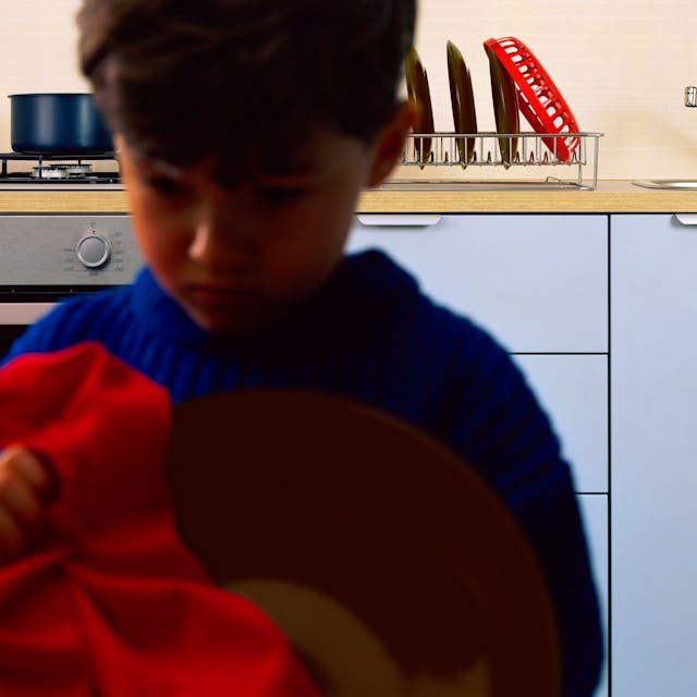 Ein Junge sitzt in einer Küche, im Hintergrund sind ein Waschbecken und Geschirr zu sehen.