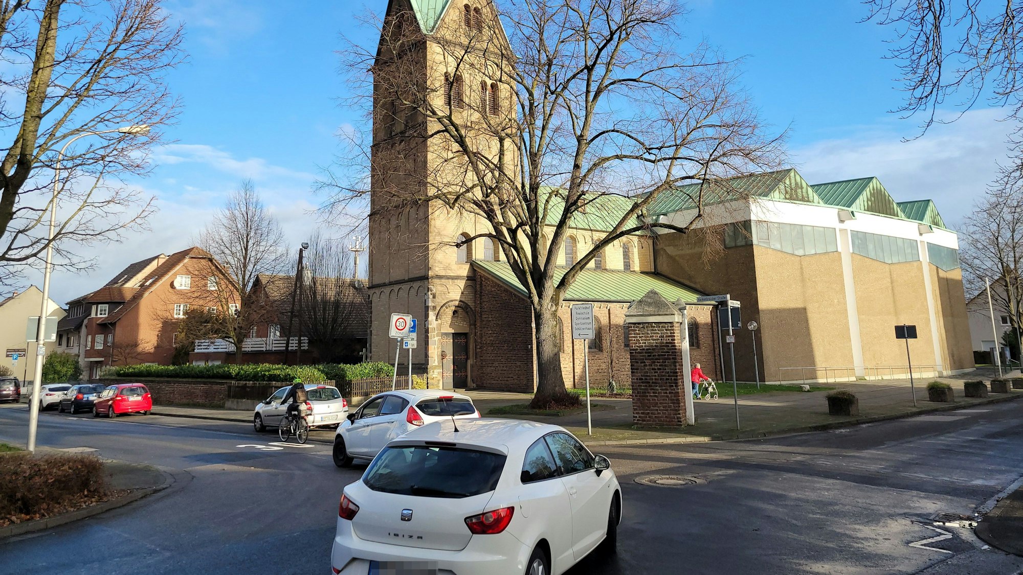 Zu sehen ist eine Kreuzung, mehrere nach links abbiegende Autos, eine Radfahrerin und im Hintergrund eine Kirche.