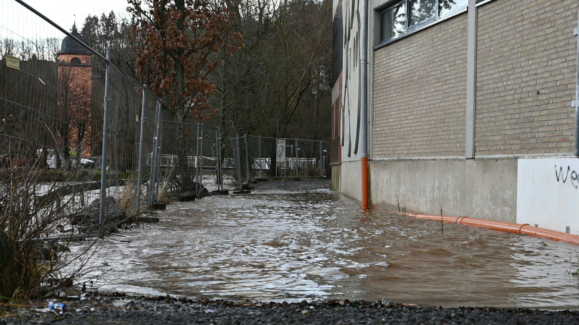 Links hinter einem Bauzaun ist die Olef in Schleiden. Der Fluss führt Hochwasser, das Wasser reicht bis ans Sturmius-Gymnasium heran.