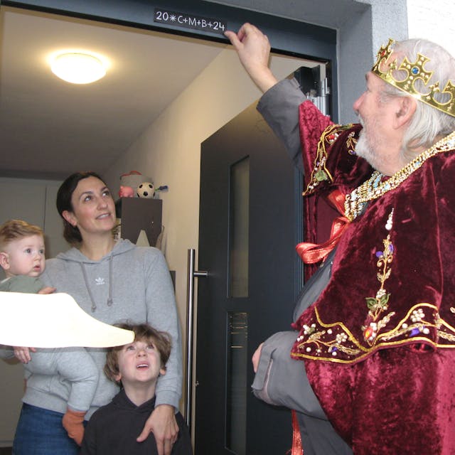 Sternsinger Wolfgang Czarnecki bringt über einer geöffneten Haustür den Segensaufkleber an, eine Frau mit einem kleinen Jungen und einem Baby auf dem Arm steht in der Tür und schaut zu.