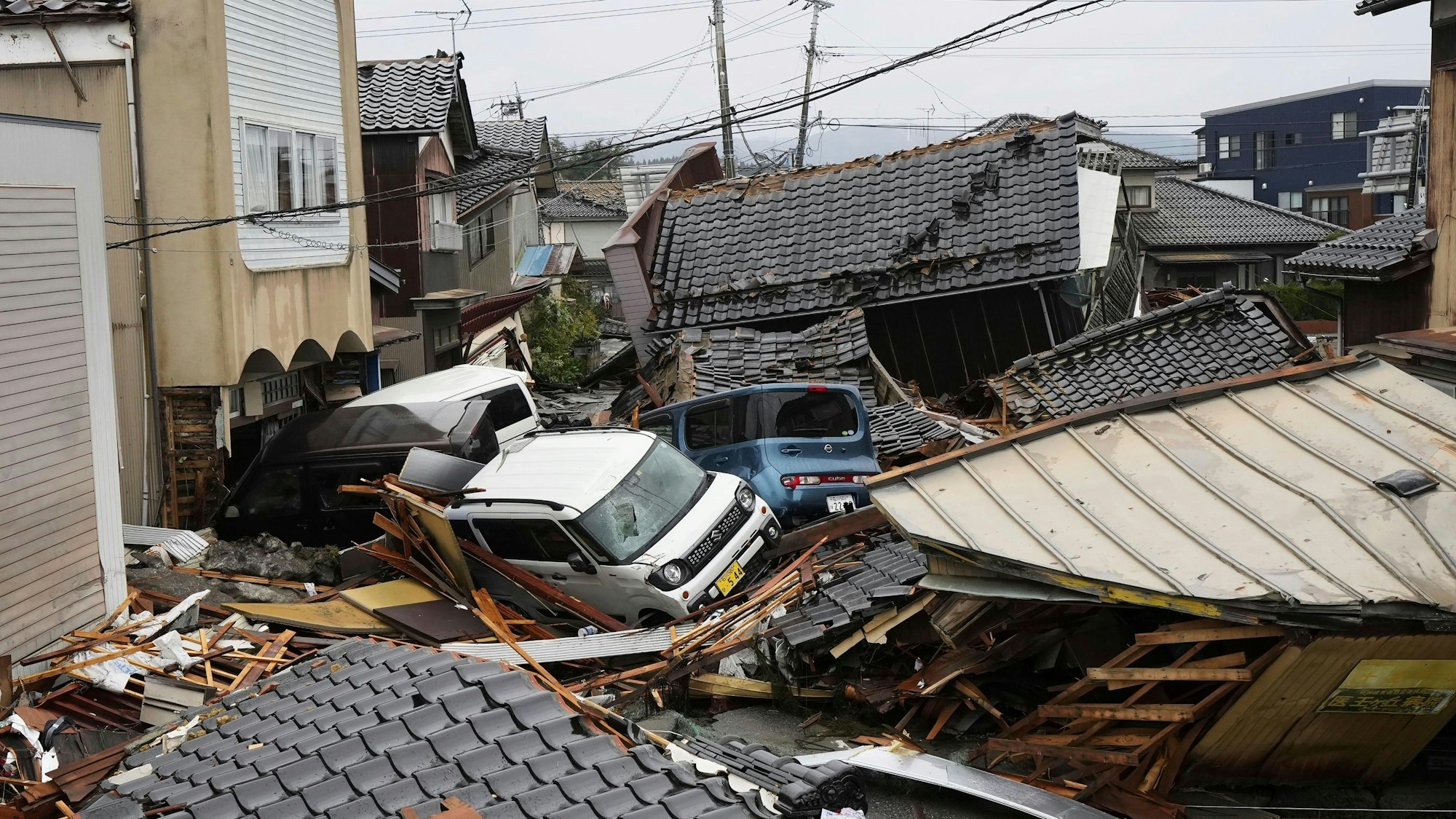 03.01.2024, Japan, Suzu: Fahrzeuge und eingestürzte Häuser sind in einem Gebiet zu sehen, das nach den Erdbeben in Suzu, Präfektur Ishikawa in Brand geraten ist. Trümmer, Schlammlawinen und aufgerissene Straßen behindern die Suche nach Überlebenden. Die Zahl der Opfer, nach dem Beben der Stärke 7,6 vom Neujahrstag, steigt weiter an. Foto: Uncredited/Kyodo News/AP/dpa +++ dpa-Bildfunk +++