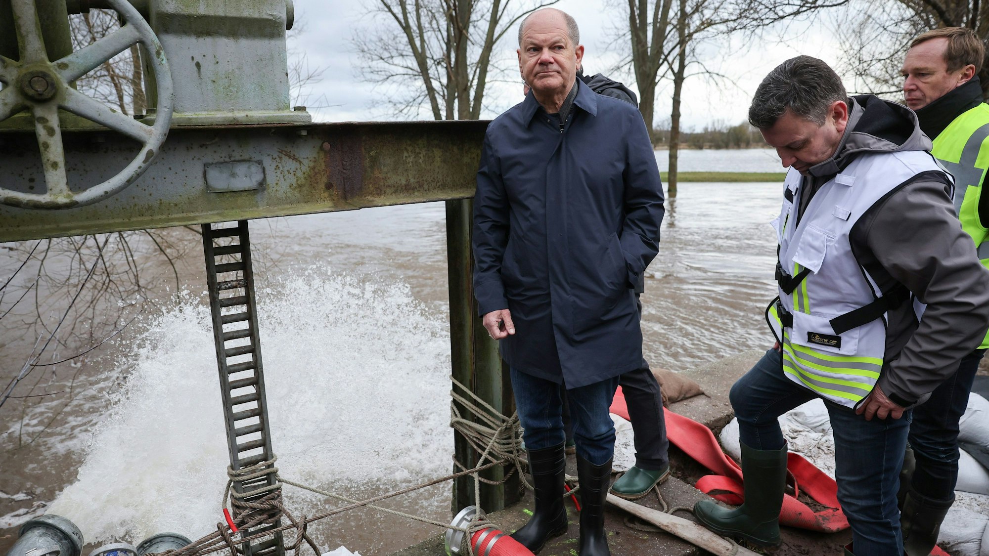 Bundeskanzler Olaf Scholz (l, SPD) steht mit André Schröder (M, CDU), Landrat Mansfeld-Südharz, an einem Wehr, an dem Schläuche abgepumptes Wasser abführen.