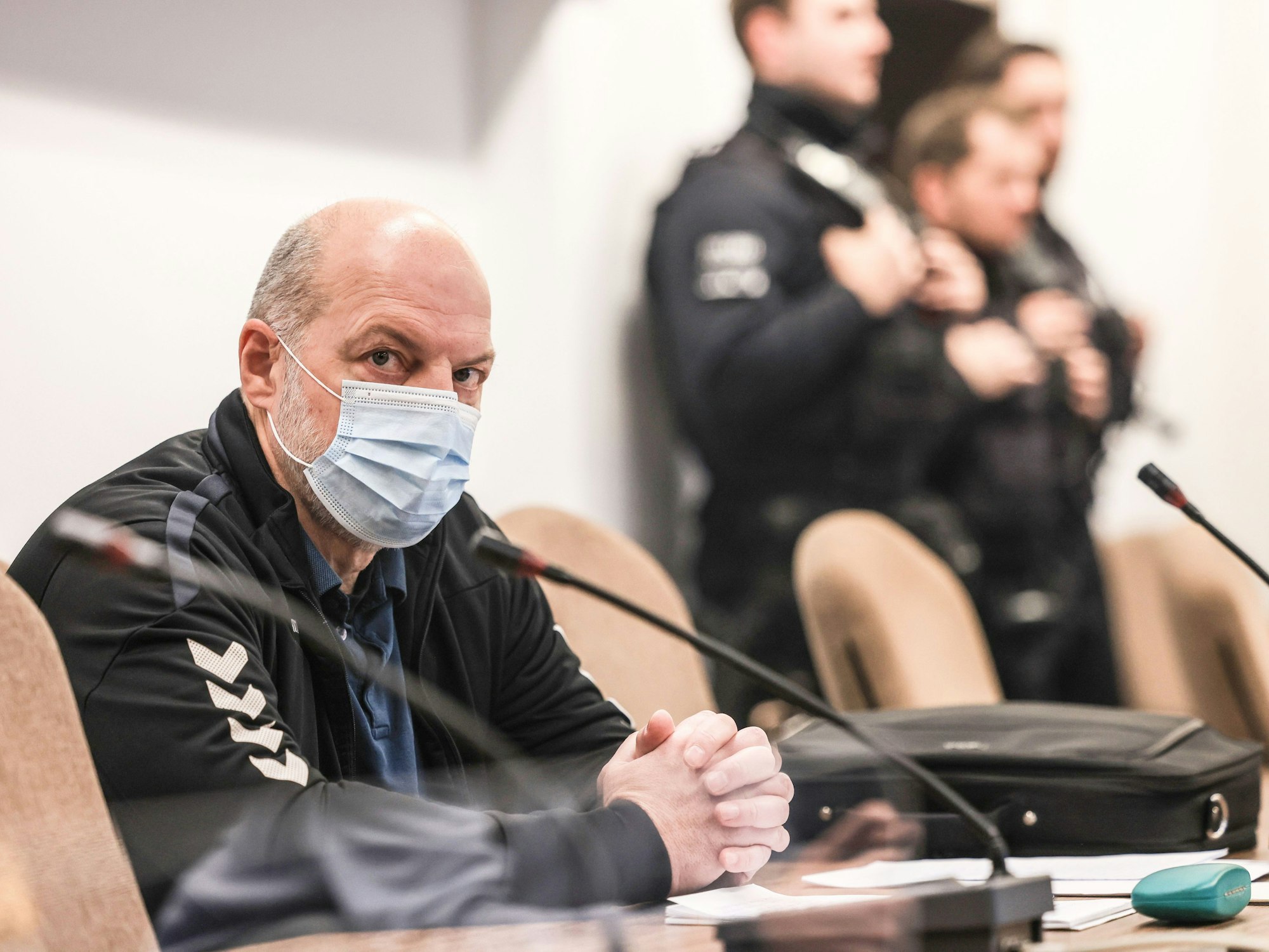 Ein Mann mit Mundschutz sitzt an einem Tisch vor einem Mikrofon. Im Hintergrund sind Polizeibeamte zu sehen.