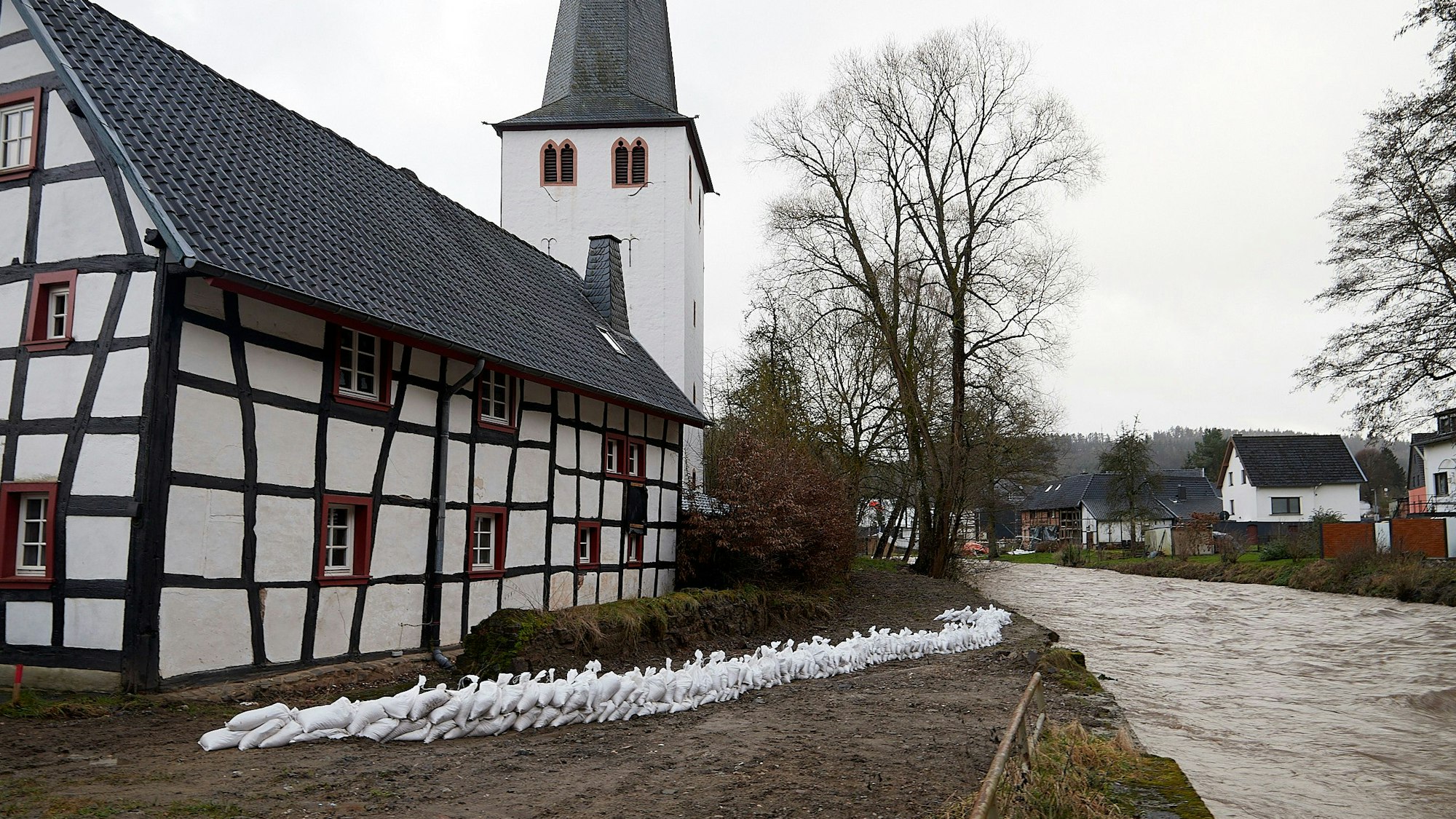 Eine Mauer aus weißen Sandsäcken ist in der Mitte des Bildes zu sehen. Rechts ist die Hochwasser führende Olef, links ein Fachwerkhaus, das durch die Säcke geschützt werden soll.