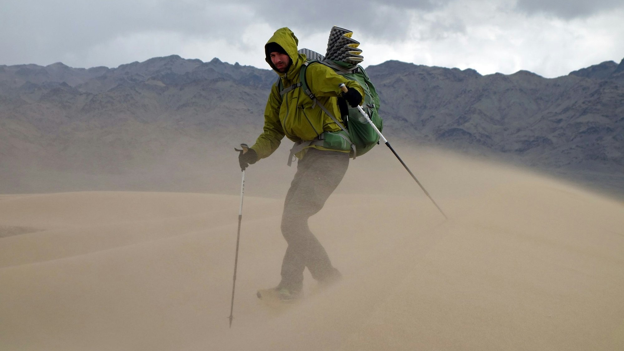 Das Bild zeigt Michael Giefer aus Lindweiler in der Wüste Gobi. Er hat einen Rucksack auf dem Rücken und geht mit Walking-Stöcken durch den vom Wind aufgewirbelten Sand einer Düne.