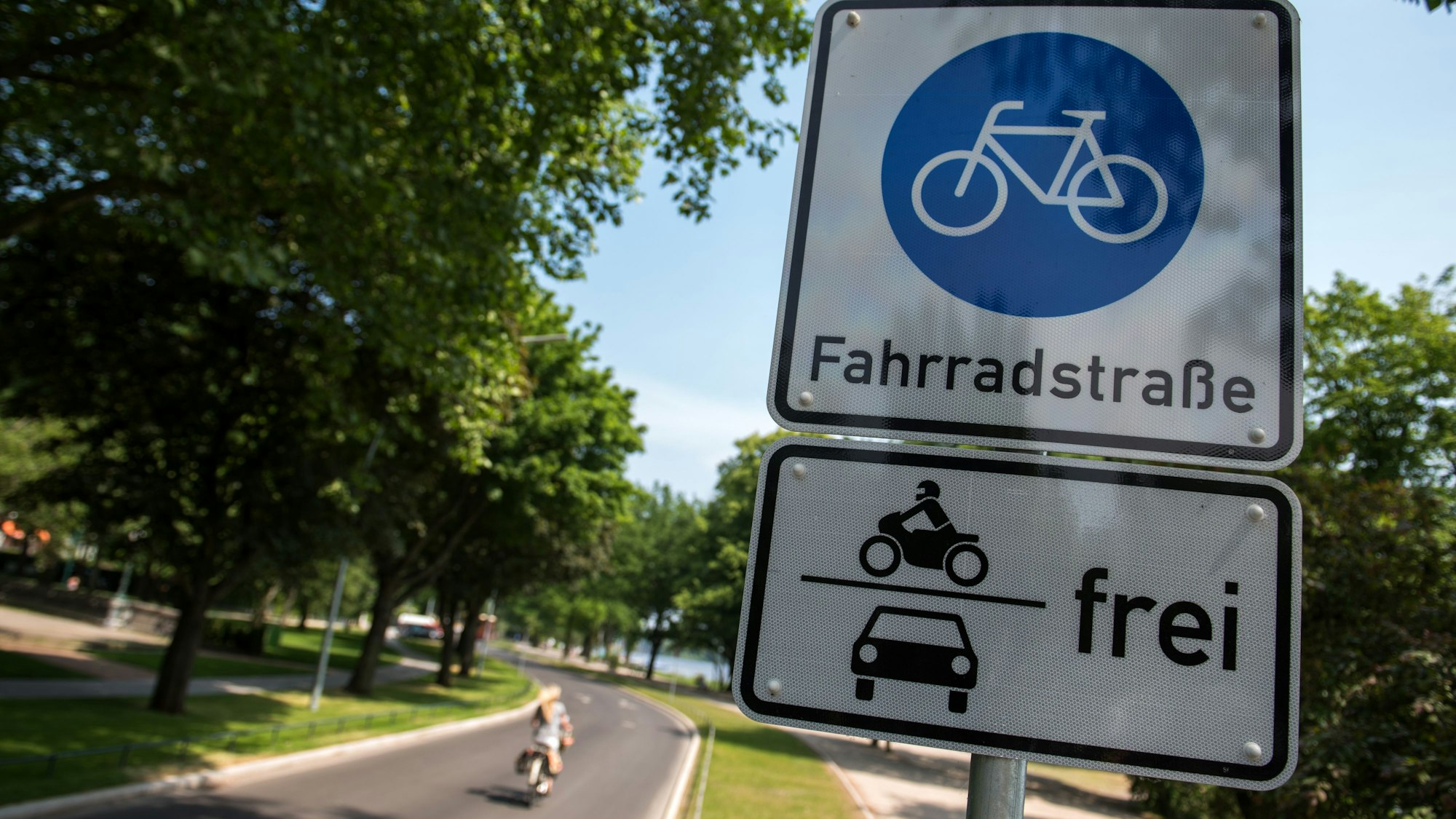 Ein Schild Fahrradstraße mit dem Zusatz Motorräder und Autos frei hängt an einer Fahrradstraße, auf der eine Radfahrerin unterwegs ist.