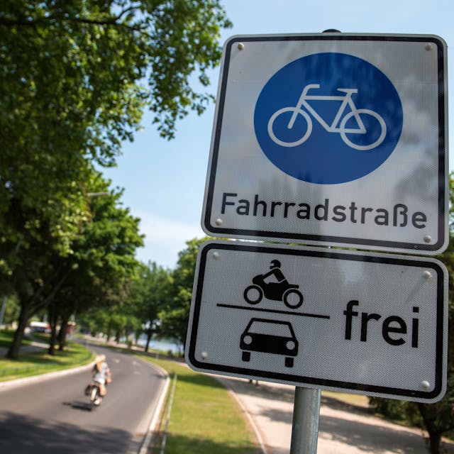 Ein Schild Fahrradstraße mit dem Zusatz Motorräder und Autos frei hängt an einer Fahrradstraße, auf der eine Radfahrerin unterwegs ist.&nbsp;