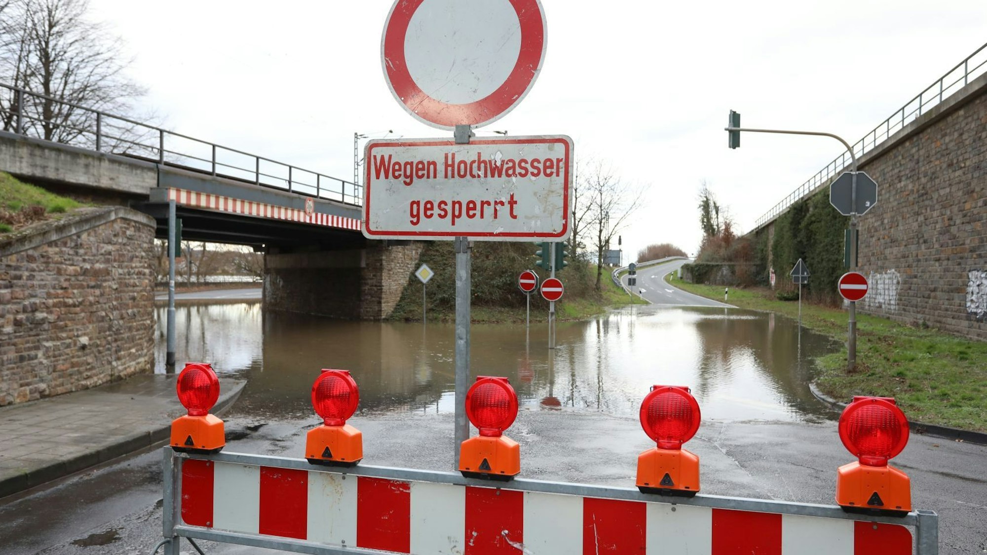 Rot-Weiße Sperren und ein Schild „Wegen Hochwasser gesperrt“ stehen vor einem überfluteten Straßenabschnitt.