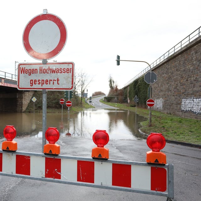 Rot-Weiße Sperren und ein Schild „Wegen Hochwasser gesperrt“ stehen vor einem überfluteten Straßenabschnitt.