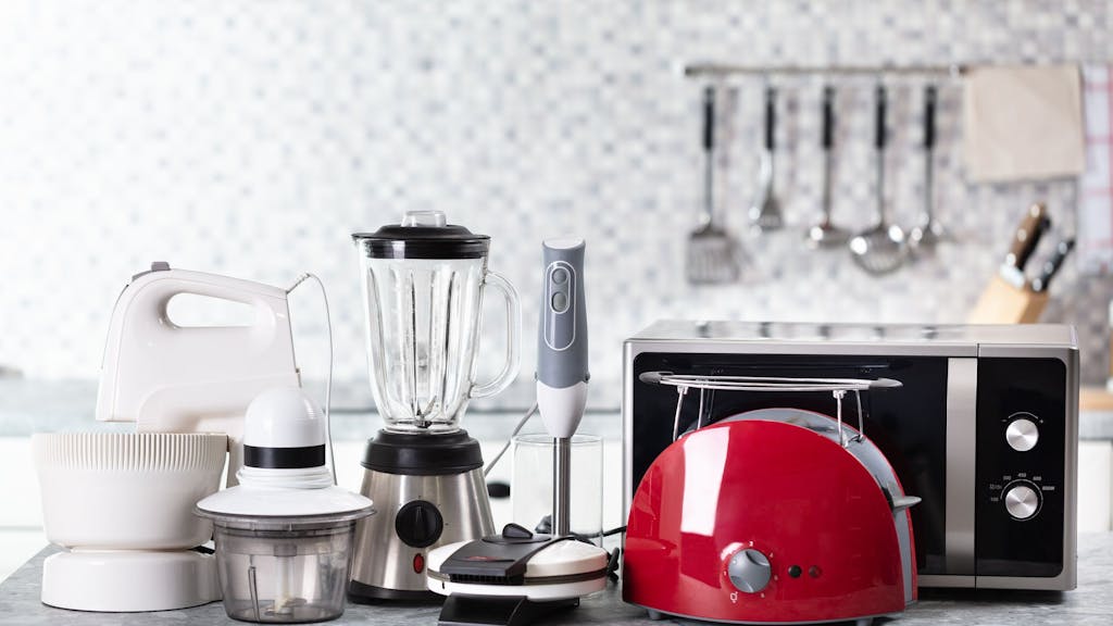 Auf einem Küchentisch stehen mehrere Küchengeräte wie Mikrowelle, Standmixer oder Pürierstab.
