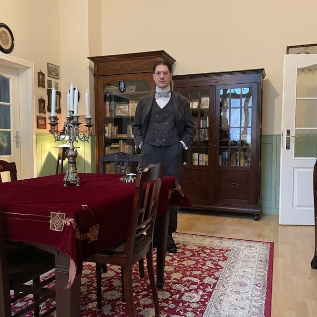 Ein Mann im altmodischen Anzug und Hemd mit Fliege steht in einem altmodisch eingerichteten Wohnzimmer.
