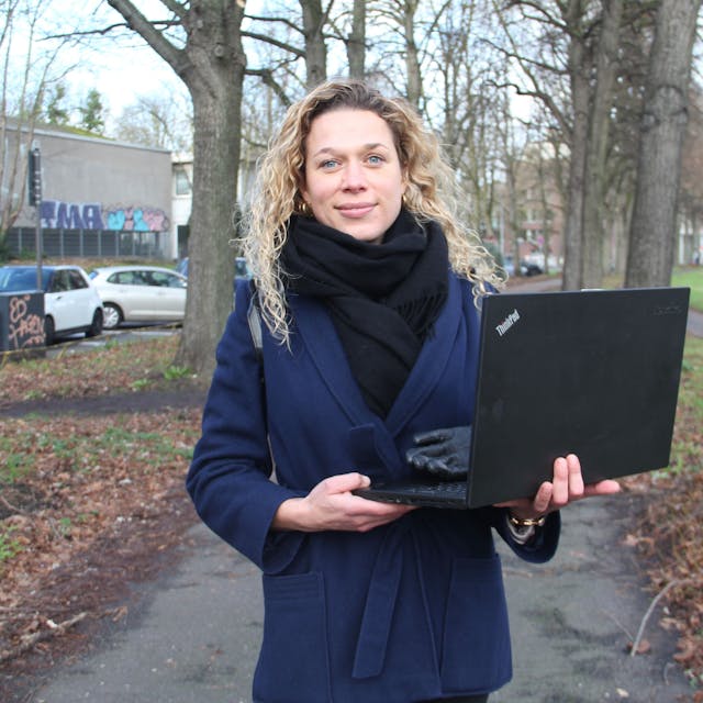 Eine blonde junge Frau steht mit einem Laptop in der Hand auf einem Spazierweg zwischen hohen Bäumen.