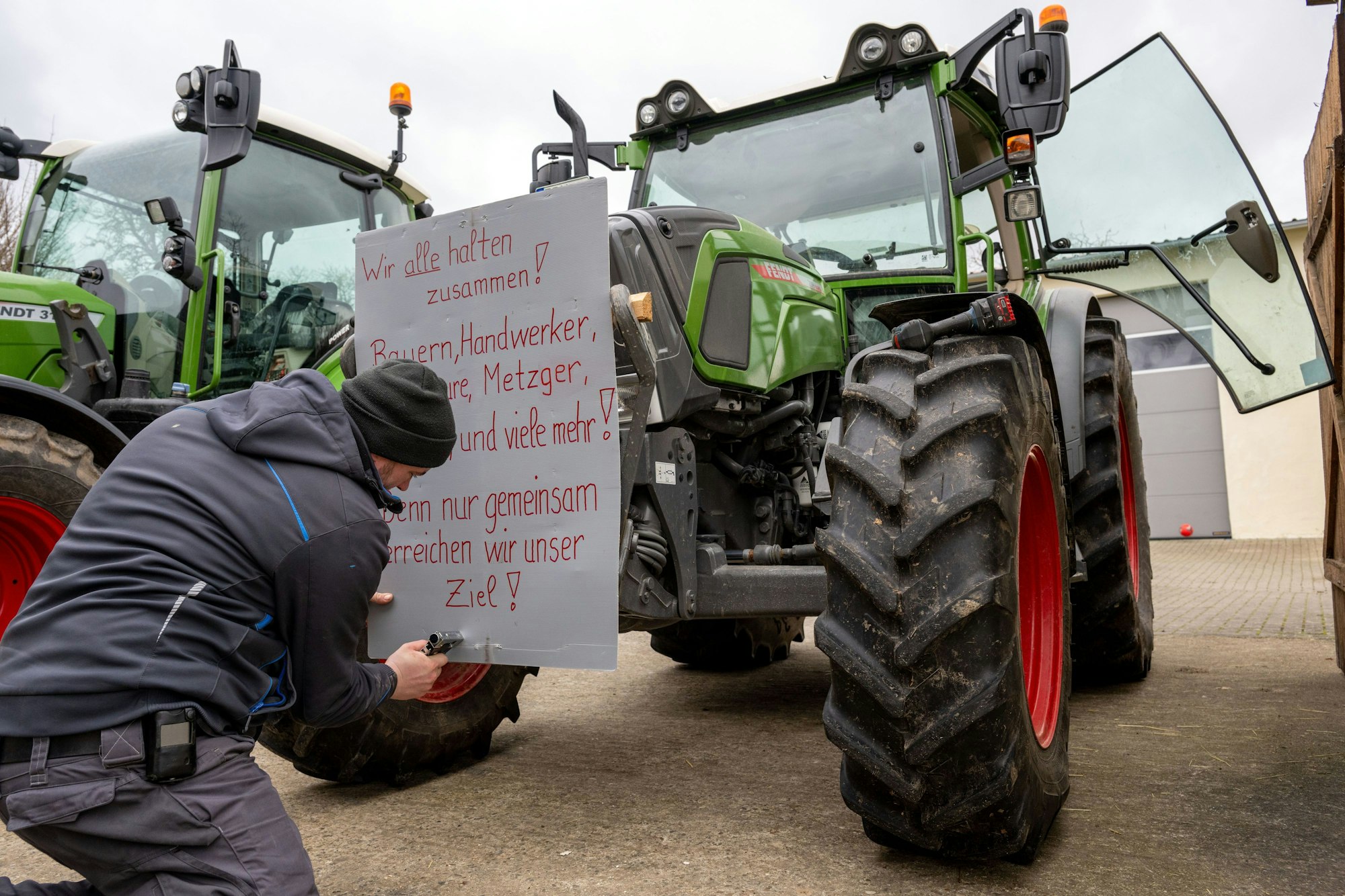 07.01.2024, Bayern, Wüstenwelsberg: Ein Mann befestigt ein Protestschild mit der Aufschrift «Wir alle halten zusammen! Bauern, Handwerker, Spediteure, Metzger, Bäcker, und viele mehr! Denn nur gemeinsam erreichen wir unser Ziel!» an einem Schlepper. Die Landwirte bereiten sich auf die anstehende Aktionswoche vor. Foto: Pia Bayer/dpa +++ dpa-Bildfunk +++