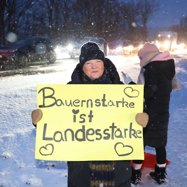 Eine Frau steht im Schneegestöber an der B51 bei Blankenheim und bekundet mit einem Transparent ihre Solidarität mit den Landwirten. Deren Demonstrationszug ist im Hintergrund zu sehen.