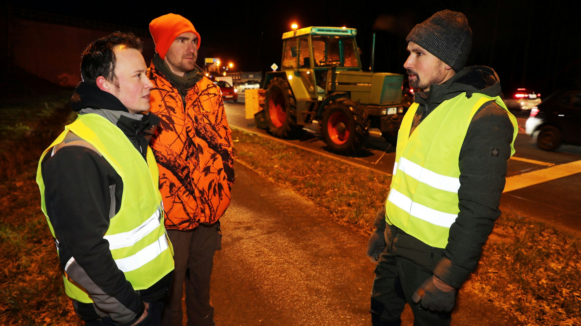 Drei protestierende Landwirte und Transportunternehmer stehen vor einer mit einem Traktor blockierten Straße in Overath.