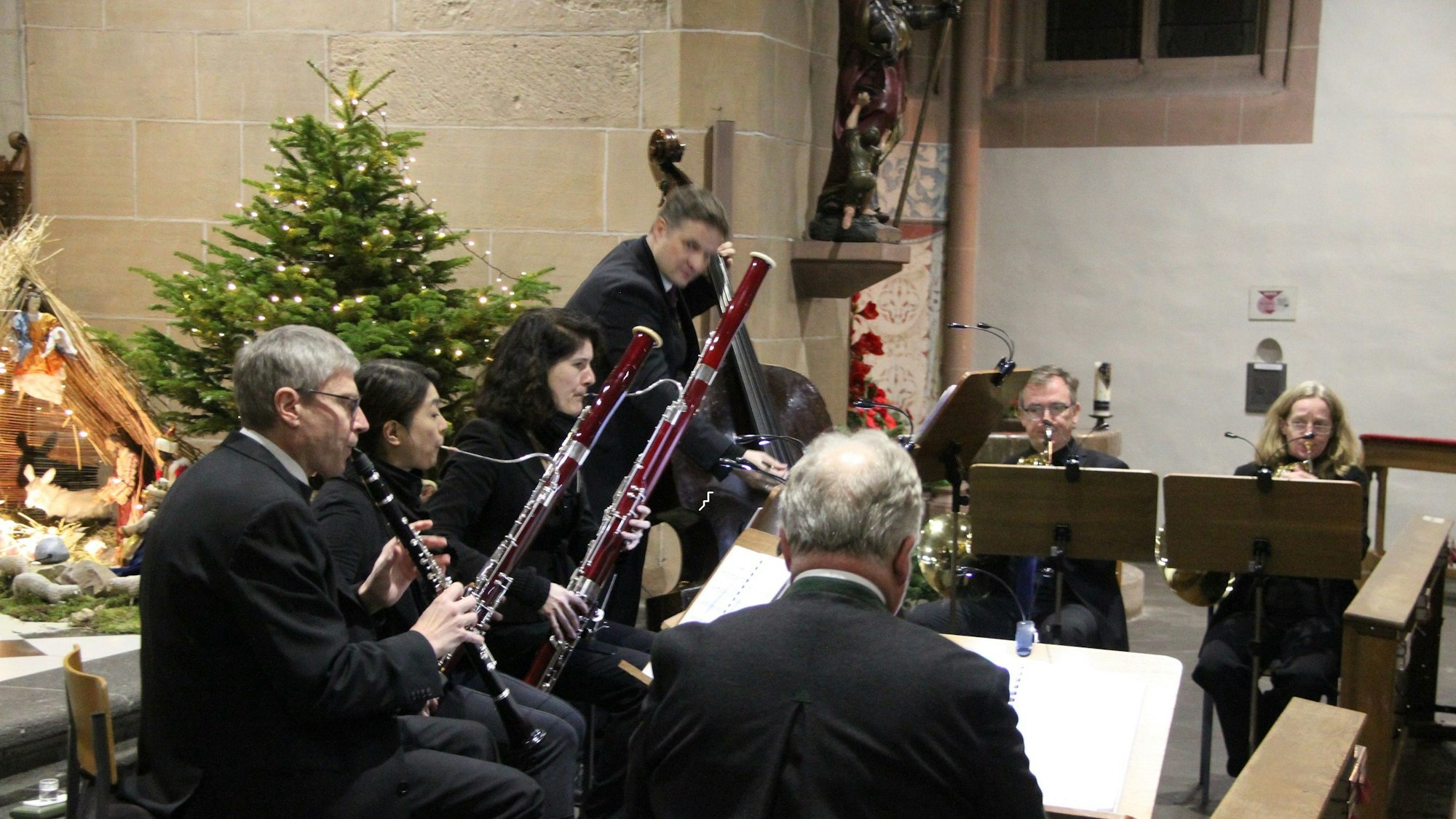 Sieben Musikerinnen und Musiker, unter anderem mit Klarinette, Fagott und Kontrabass, spielen in der Martinskirche. Vom Bassisten abgesehen, sitzen sie. Im Hintergrund stehen die Weihnachtskrippe und ein Weihnachtsbaum.