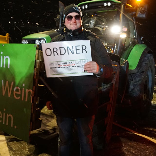 Landwirt Sebastian Bützler steht mit einem „Ordner“-Schild neben seinem Trecker auf dem Parkplatz in Breitenbenden. Auf dem Banner seines Treckers steht: „Kein Bauer, kein Bier, Schnaps, Wein – das wäre gemein!“