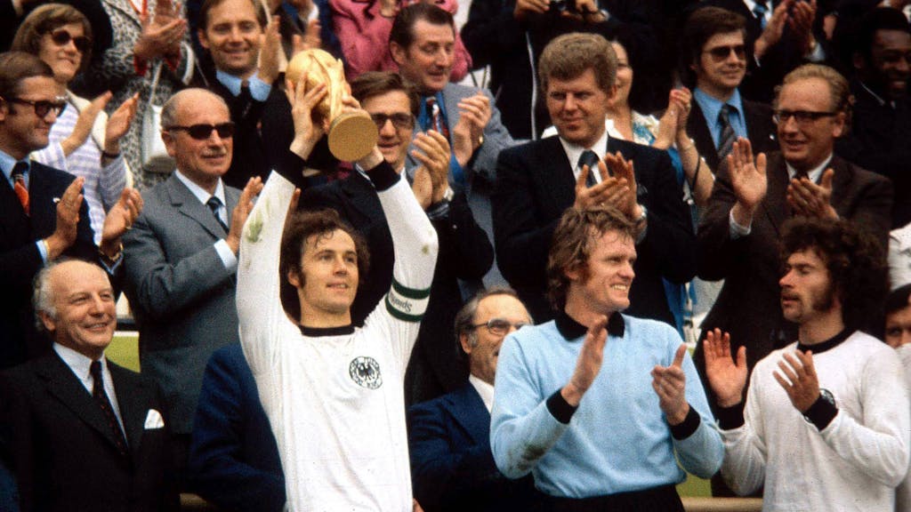 Franz Beckenbauer reckt den WM-Pokal in die Luft. Sepp Maier und Paul Breitner applaudieren.