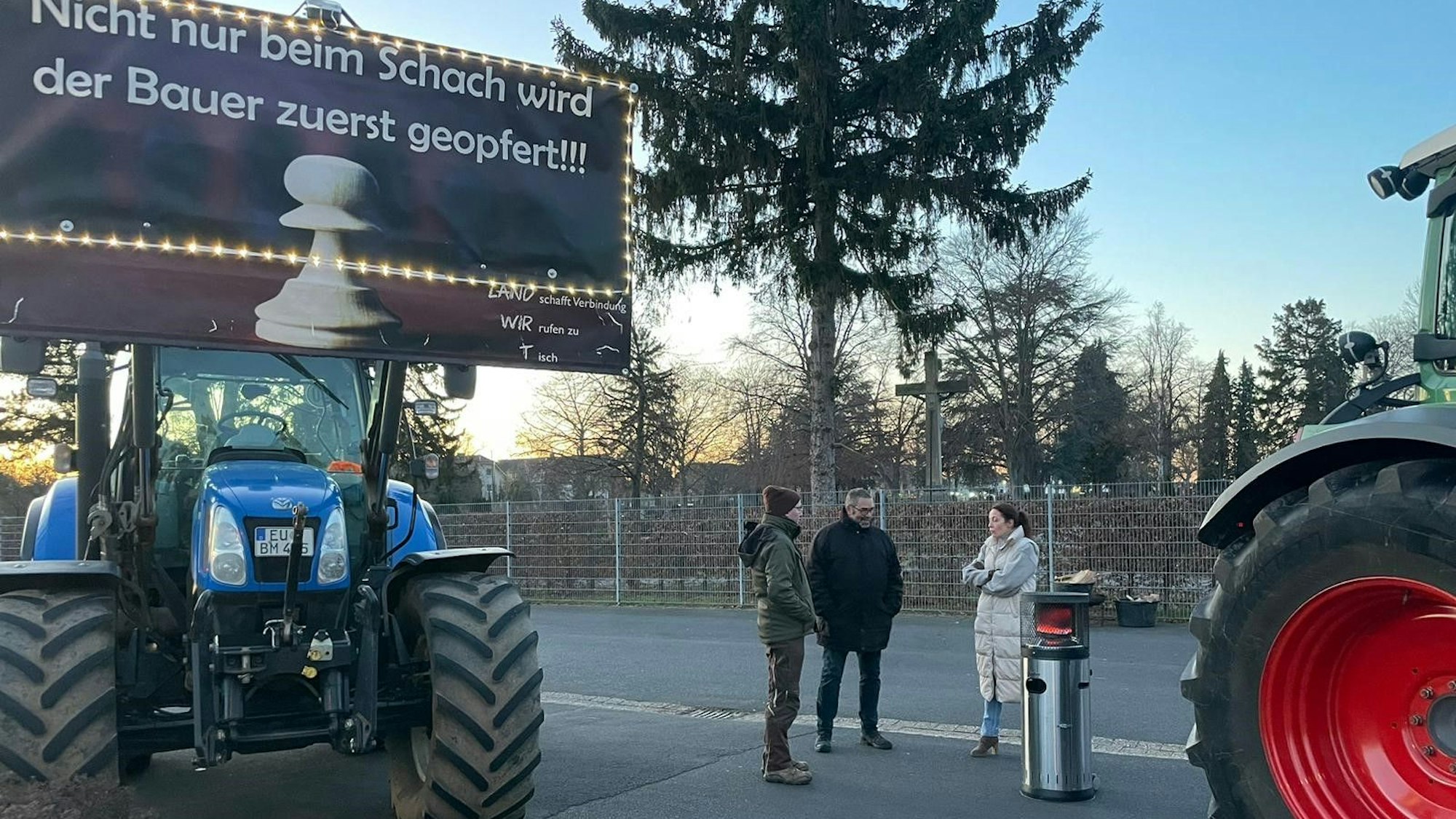 Zwei Männer und eine Frau stehen an einem Heizpilz. Neben ihnen stehen zwei Traktoren mit Protestplakaten.