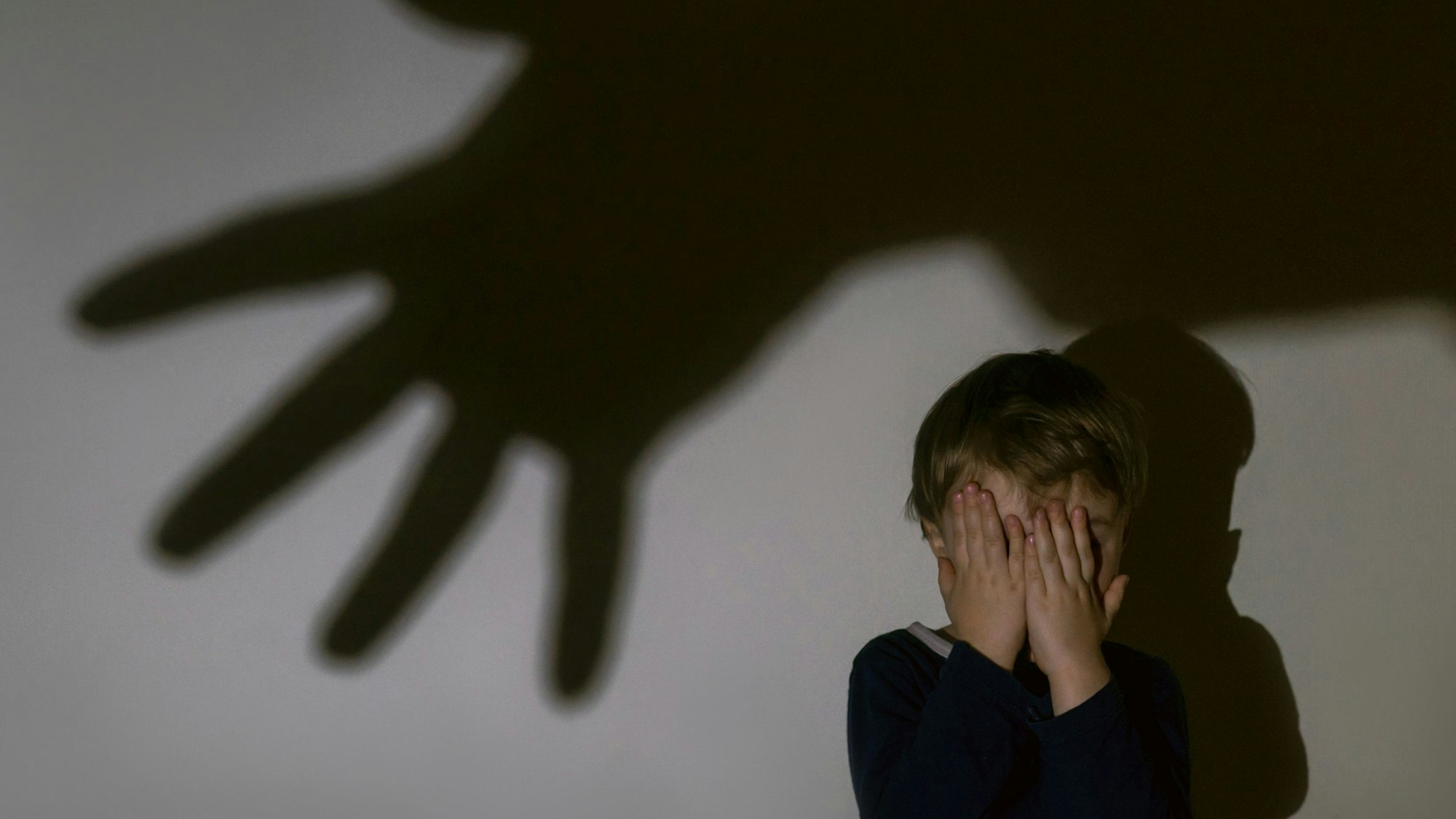Ein kleiner Junge sitzt vor einer Wand und hält vor Schrecken seine Hände vors Gesicht. An der Wand ist ein großer Schatten einer ausgestreckten Hand zu sehen.
