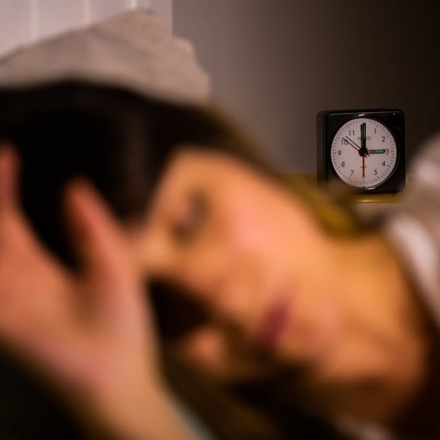 Eine Frau schläft neben einem Wecker, der auf drei Uhr zeigt.