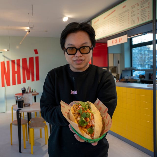 Ein Mann steht in einem Restaurant und hält ein Sandwich in die Kamera.