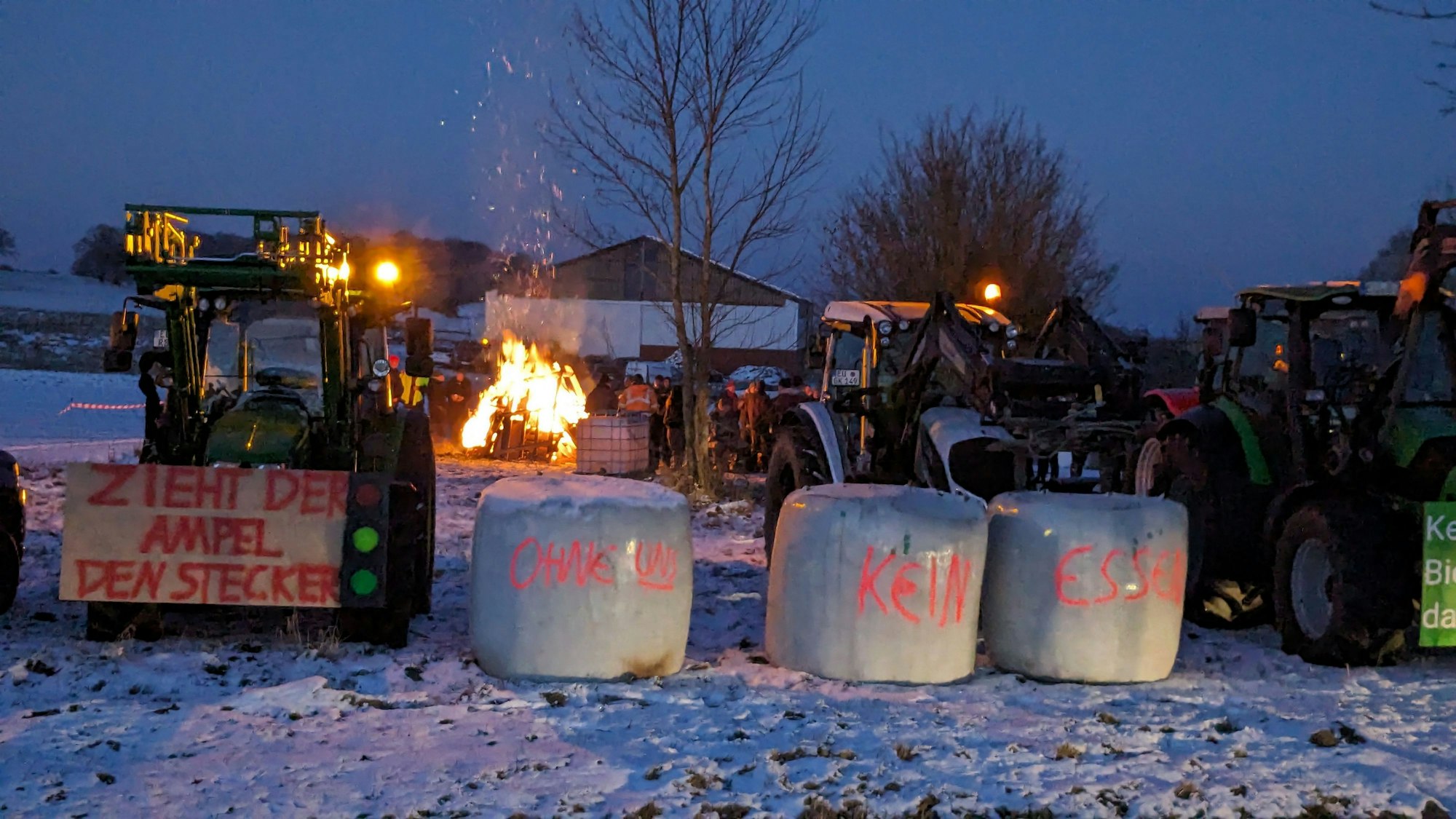 Ein Traktor und Strohballen mit Protest-Aufschriften stehen vor einem Feuer.