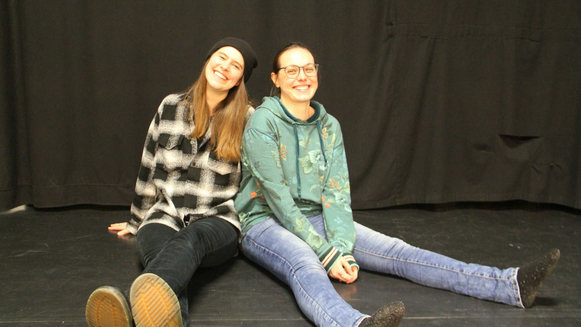 Zwei junge Frauen sitzen mit ausgestreckten Beinen auf einer Bühne.