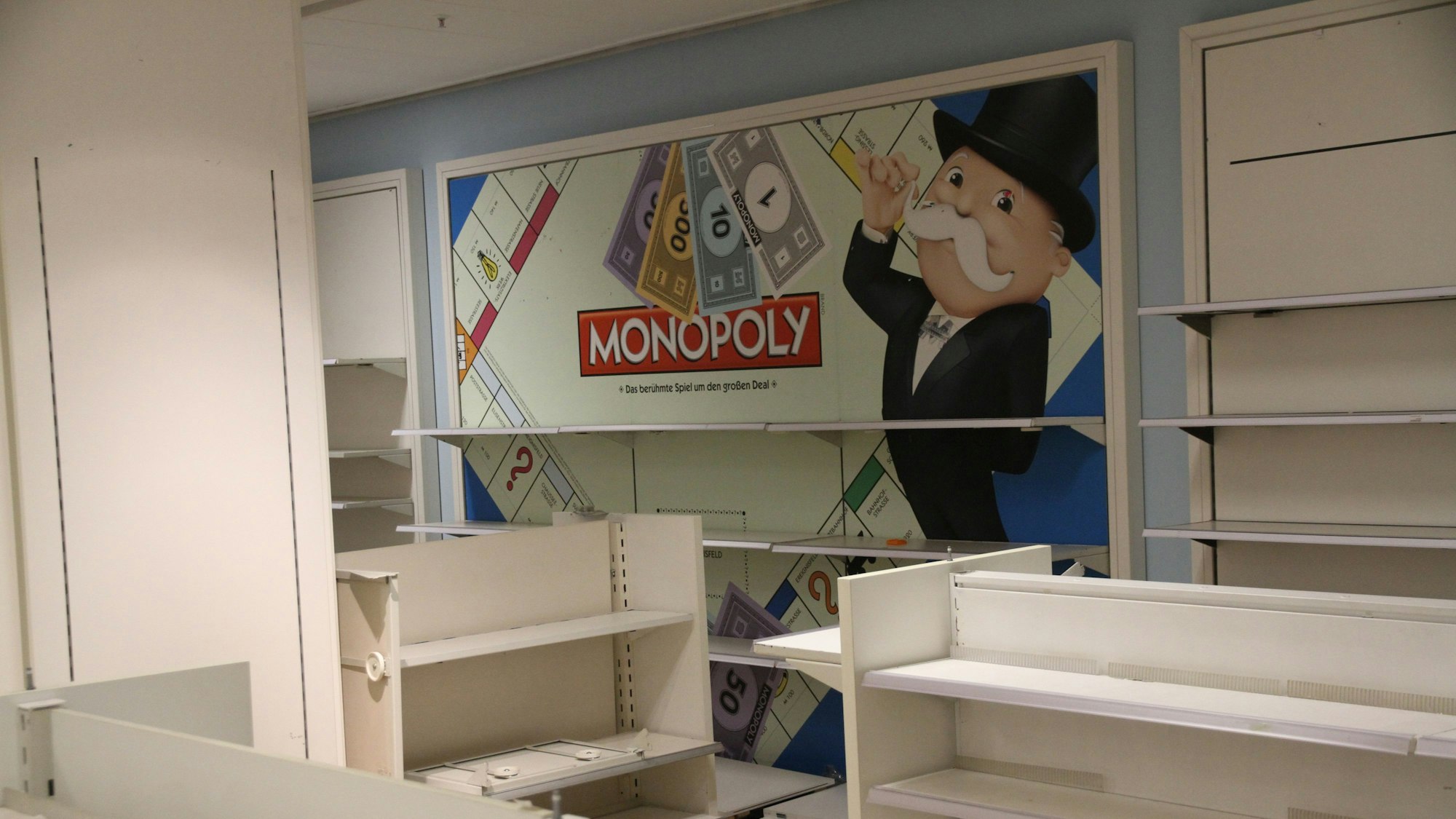 Ein Männchen mit dunklem Anzug und Schnauzbart wirbt an einer Wand der Spielwarenabteilung für das Spiel Monopoly.