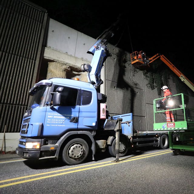Ein blauer Lastwagen mit Anhänger und ein grüner Hubsteiger stehen nachts neben einer hohen Betonwand. Die Bundesstraße ist gesperrt.