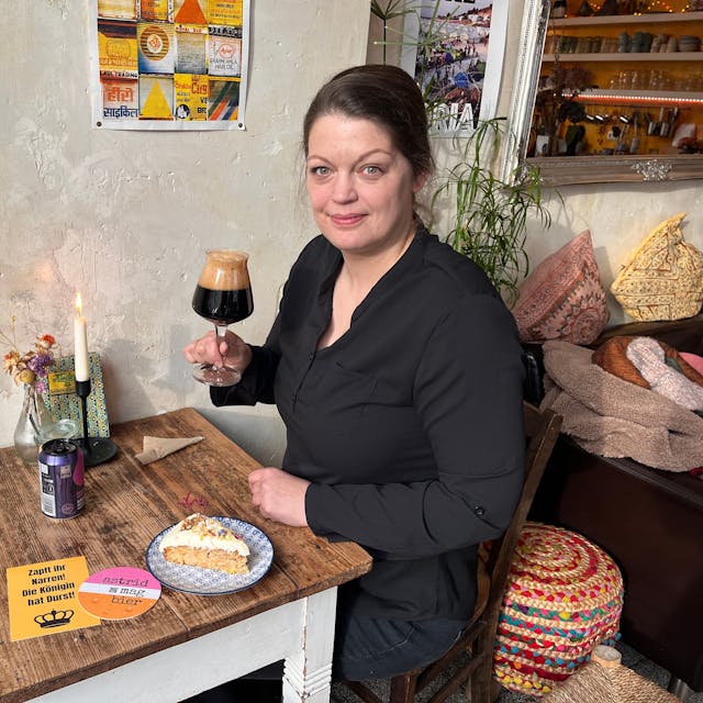 Eine Frau hat sitzt an einem Café-Tisch mit einem Glas Bier in der Hand und einem Stück Kuchen.