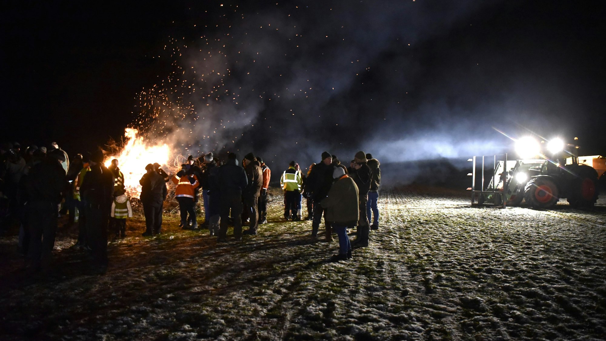 Eine Gruppe Menschen steht im Dunkeln an einem großen Feuer, rechts im Bild ist ein Traktor mit angeschaltetem Licht zu sehen.