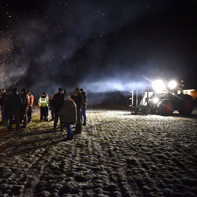 Eine Gruppe Menschen steht im Dunkeln an einem großen Feuer, rechts im Bild ist ein Traktor mit angeschaltetem Licht zu sehen.