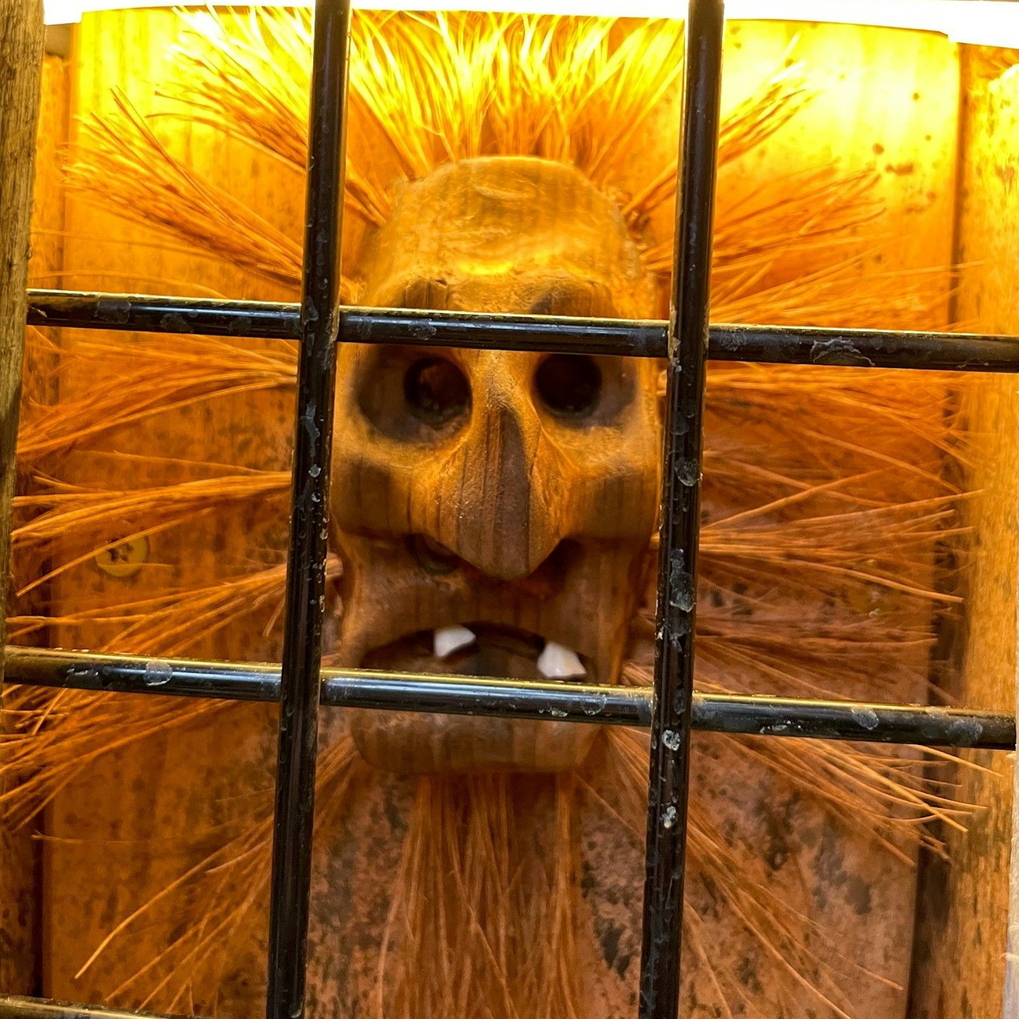Eine Maske aus Holz mit Stroh-Haaren, die in alle Richtungen zeigen, ist an einer Wand ausgestellt.