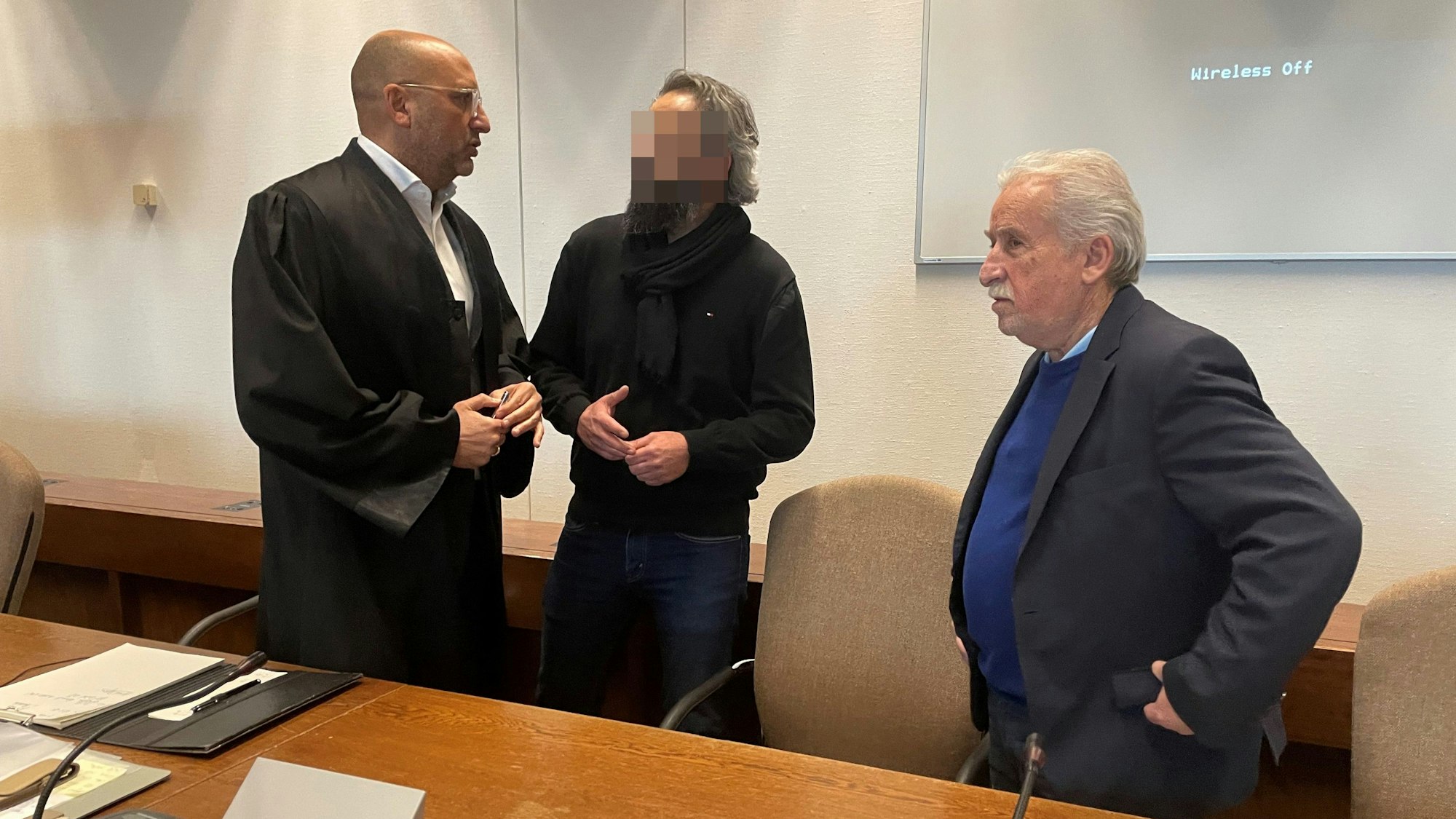 Der Angeklagte mit Verteidiger Abdou Gabbar und Dolmetscher Bahadir Aksungur im Gerichtssaal, die Männer stehen an einem Tisch