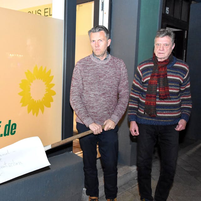 Auf dem Foto sind die Elsdorfer Grünen-Politiker Michael Broich und Hartmut Bergheim vor dem Büro ihrer Fraktion zu sehen. Broich hält die Harke in den Händen.