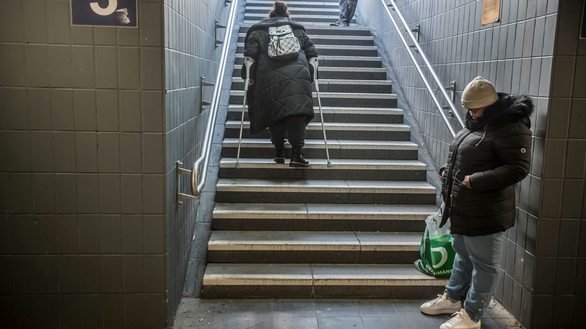 Eine Frau mit Krücken geht eine Treppe im Bahnhof Leverkusen-Mitte hoch. Eine andere Frau wartet im Treppenschacht.