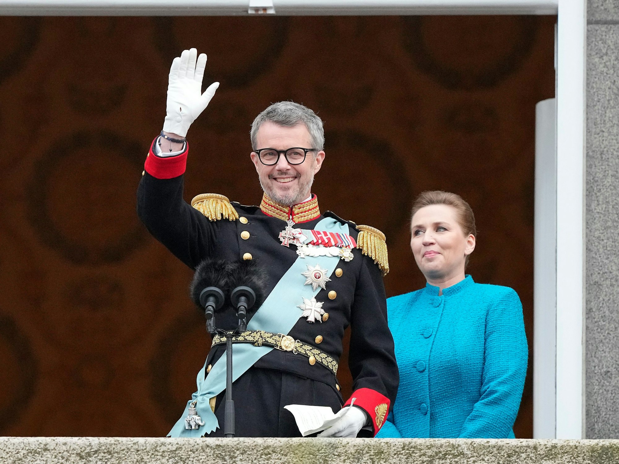 König Frederik X. von Dänemark winkt vom Balkon des Schlosses Christiansborg, während die dänische Ministerpräsidentin Mette Frederiksen zusieht.